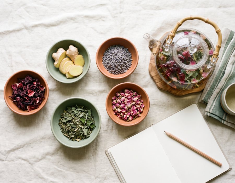 Flat-lay of herbal tea ingredients, a glass teapot and a notebook on a cream linen surface